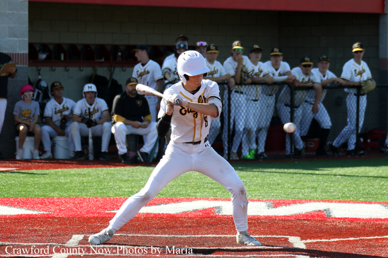 Baseball batter in a white uniform with a helmet swings at a pitch near home plate, teammates watching from the dugout in the background.