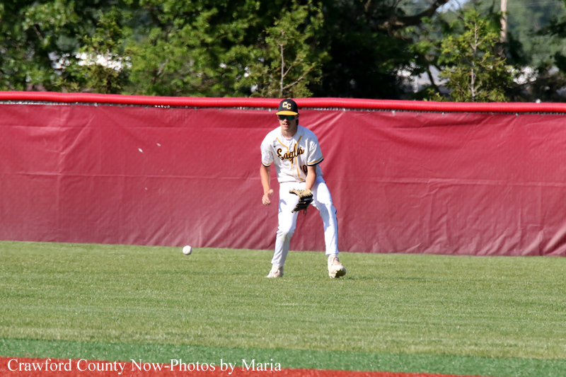 Baseball fielder in white uniform and black cap fields a ground ball on a grassy outfield with a red wall behind him.