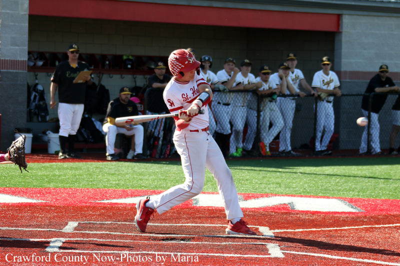 Baseball batter in a red helmet and white uniform swings at a pitch at home plate, teammates watching from the dugout in the background.