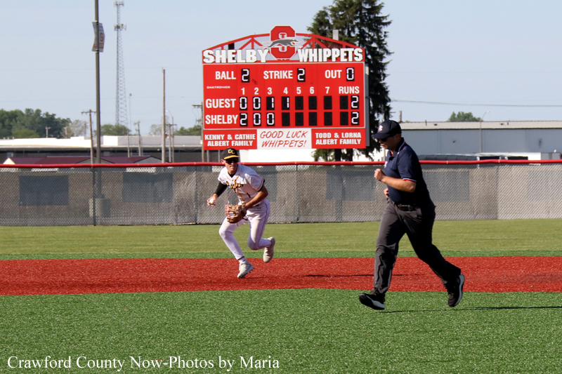 Baseball scene: a player in white uniform runs on the infield as an umpire in dark clothing follows; a large red scoreboard behind them shows 2-2 counts.