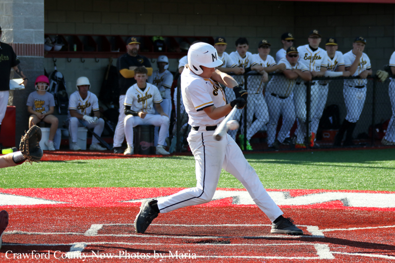 Baseball batter in a white uniform swinging at a pitch near home plate, teammates watching from the dugout in the background.