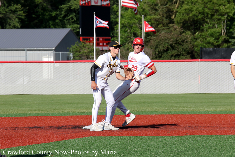 Two baseball players near a base on a sunny field: a fielder in a white uniform with a black cap stands ready as a runner in white with a red helmet approaches the base.