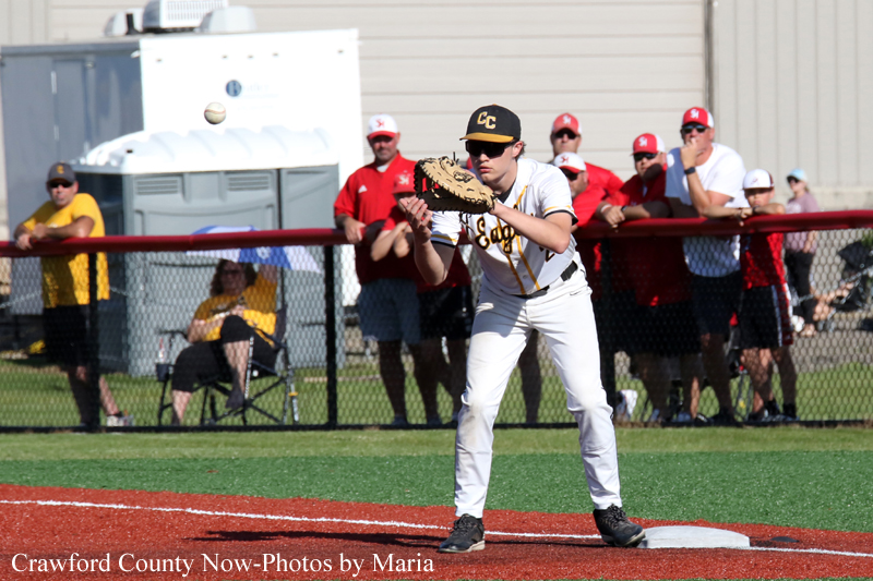 Baseball infielder in a white uniform with a CC cap reaches for a ball near first base as spectators watch behind a fence.