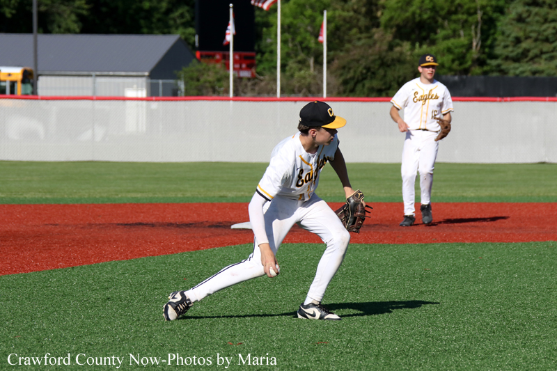 Baseball pitcher in a white and yellow Eagles uniform lunges to field a ground ball on artificial turf; teammate watches from the infield in the background.