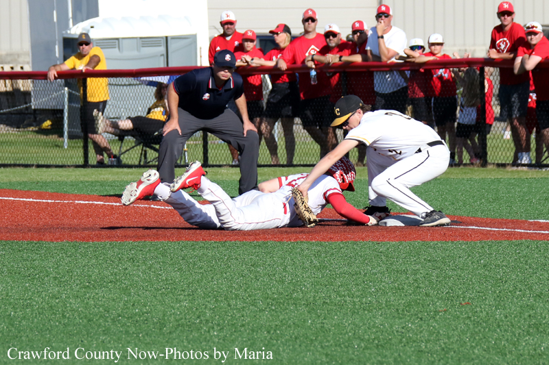 Baseball play at home plate: a runner slides in as a fielder reaches to tag him, with the umpire watching and fans behind the fence.