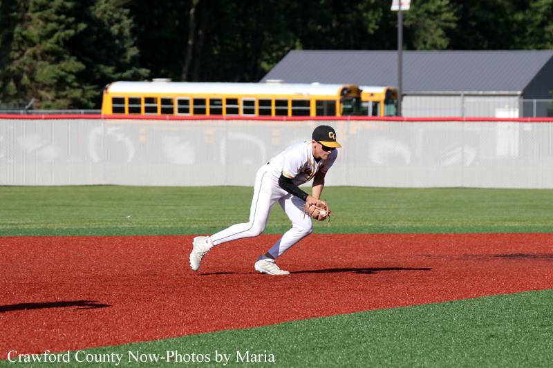 Baseball infielder in a white uniform bends to field a ground ball on the red infield dirt, glove ready, with a yellow school bus and fence in the background.