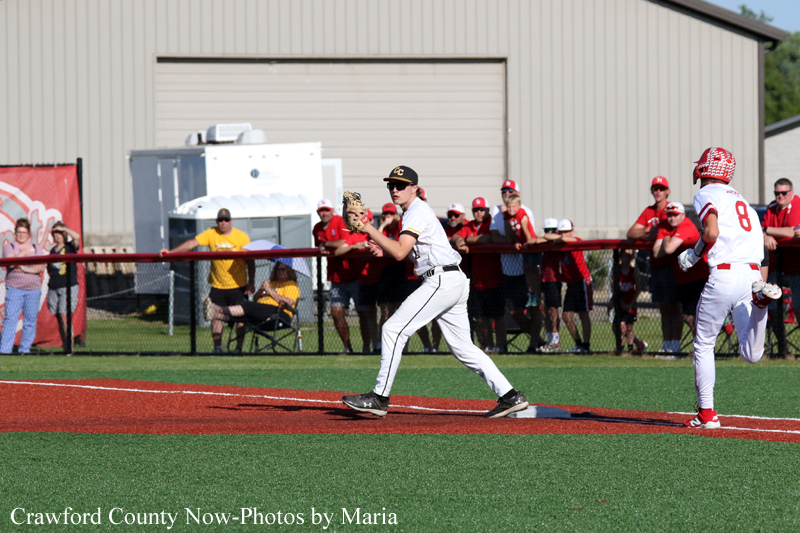 Baseball pitcher in white uniform winds up on the mound as a baserunner in white and red runs past the on-deck area, with a crowd behind a fence in the background.
