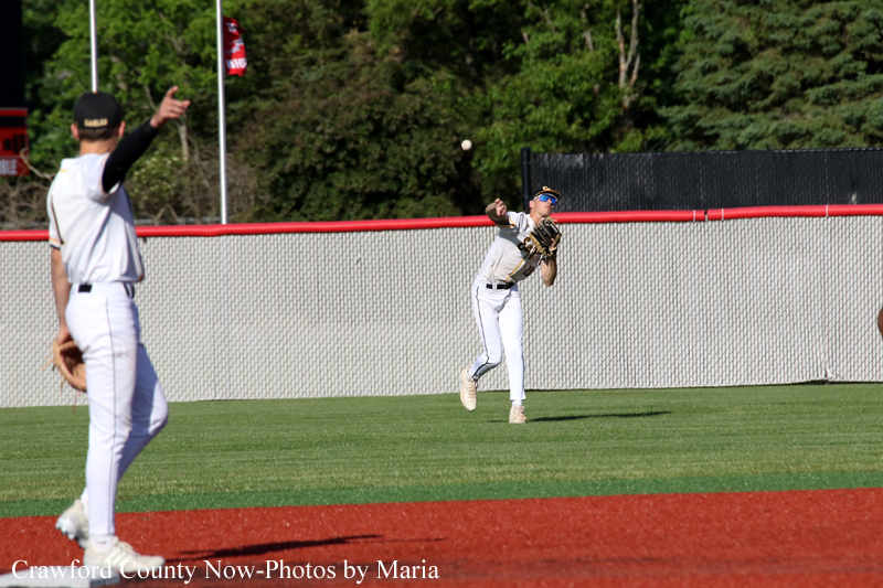 Baseball game: pitcher in white throws a pitch while a fielder watches in the foreground on a green field with a chain-link fence behind.