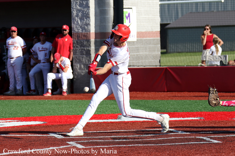 Baseball batter in white and red uniform swings at a pitch near home plate, with teammates watching in the dugout in the background.