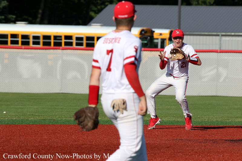 Two baseball players in white and red uniforms on a baseball field; one in the foreground with a glove watches the other ready to catch near the infield dirt, with a yellow school bus in the background.