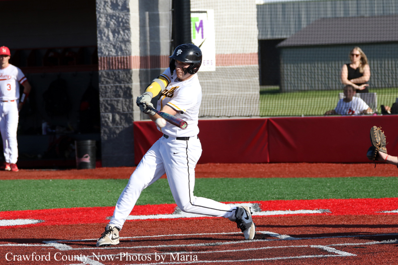 Baseball batter in a white uniform and black helmet swings at the ball on a red infield, with spectators behind the fence.