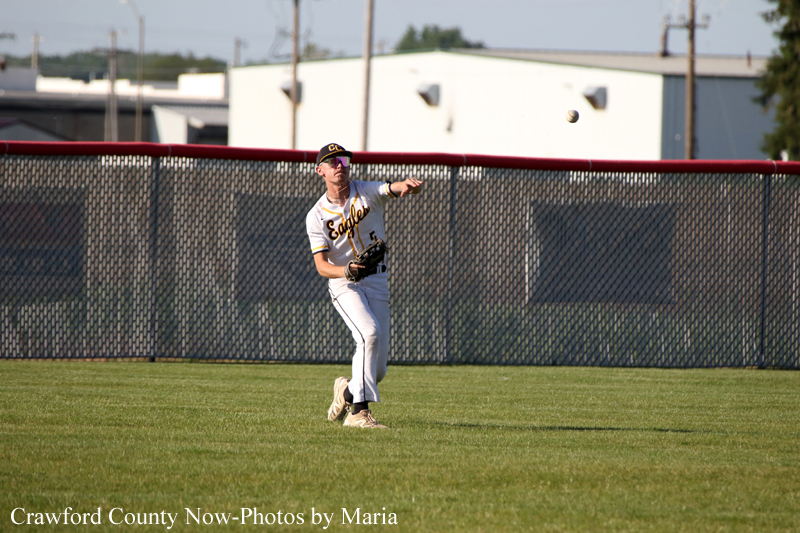Baseball player in white uniform throws a ball on a grassy field with a chain-link fence behind him.