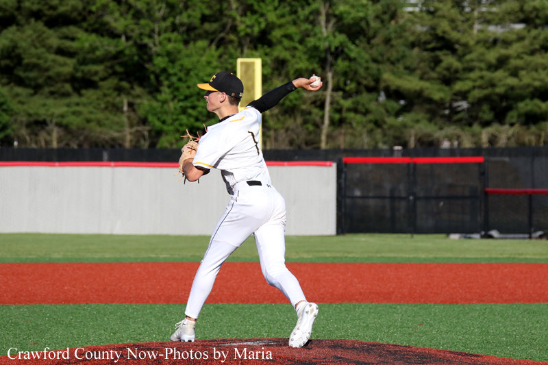 Baseball pitcher in a white uniform mid-throw on the mound with a green field and fence behind.