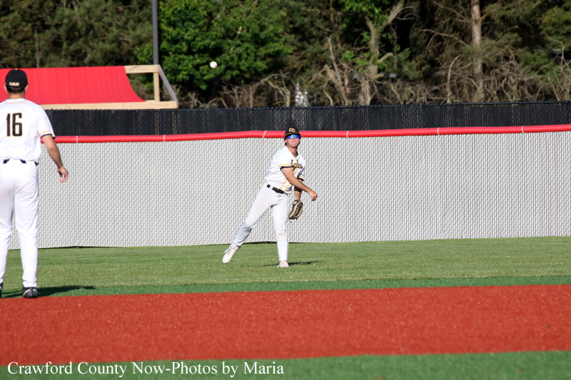 Baseball pitcher in white uniform on the mound ready to throw, fielder on the left, green field behind a chain-link fence.