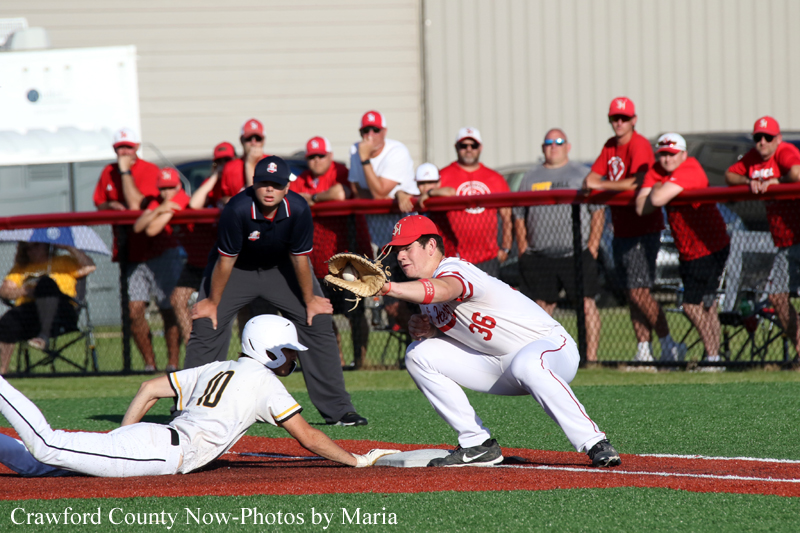 Baseball action at the base: a defender in white and red dives with his glove toward a sliding baserunner in white and gold nearby the bag; crowd watches in red shirts behind the fence.