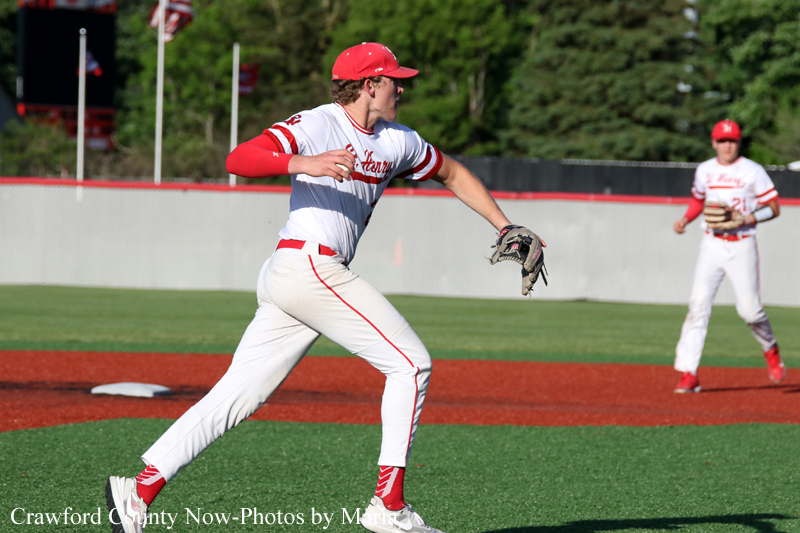 Baseball pitcher in white and red uniform winds up to throw on a green field with a teammate in the background.