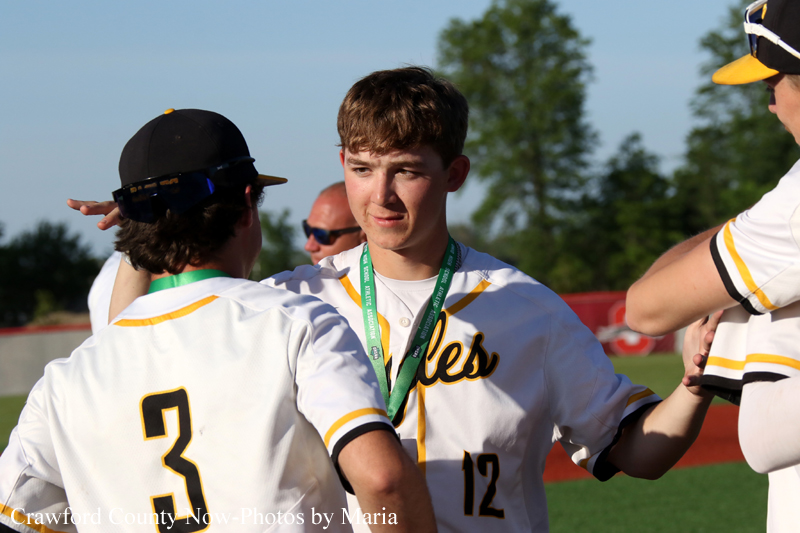 Two young baseball players in white and yellow uniforms celebrate with a high-five on a sunny field, a medal hanging from the neck of the center player.