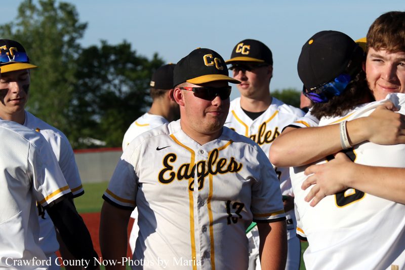 Baseball players in white Eagles uniforms with black and gold caps stand together on the field, one smiling center stage as teammates hug nearby.