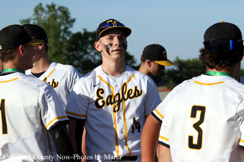 Baseball players in white and gold Eagles uniforms stand in a circle on the field; center player with number 14 and face paint looks ahead.