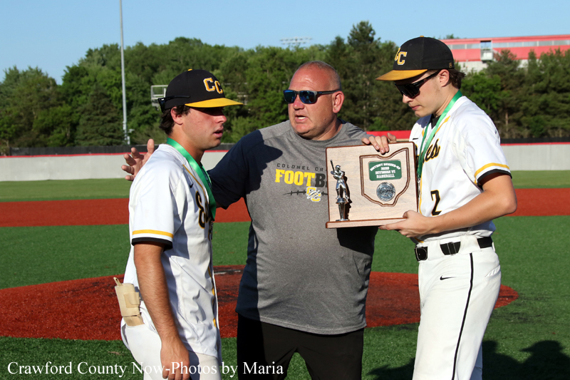 Coach hands a trophy plaque to two baseball players on a sunny field after a game, as they stand in uniform.