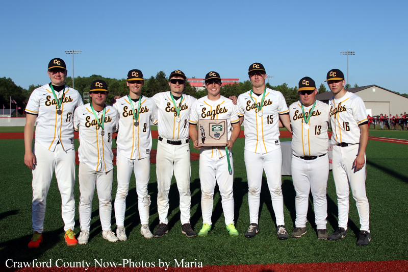 Group of nine baseball players in white and green Eagles uniforms pose on a field, one holding a trophy plaque in the center.