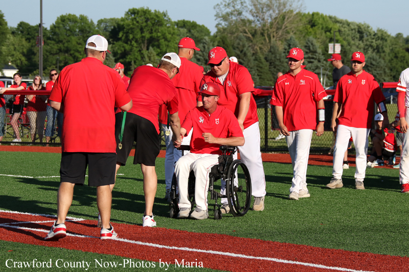 A group of baseball players in red shirts on a field; a man in a wheelchair shakes hands with a teammate while others look on.