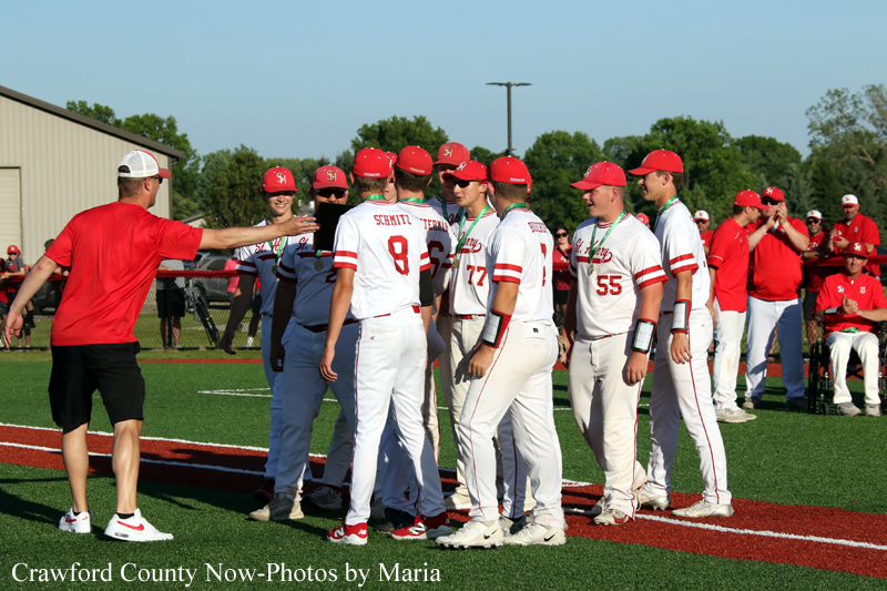 Baseball coach extends a hand to a group of players in white uniforms with red caps on the field in a team huddle.