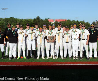 Group photo of a baseball team in white and green uniforms on a field, posing with a trophy.