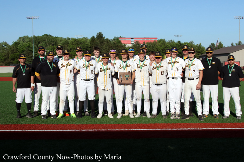 Group photo of a baseball team in white and green uniforms on a field, posing with a trophy.