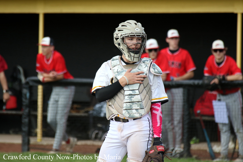 Baseball catcher in white gear with helmet and chest protector stands on the field, hand over heart, with teammates in red uniforms behind him.
