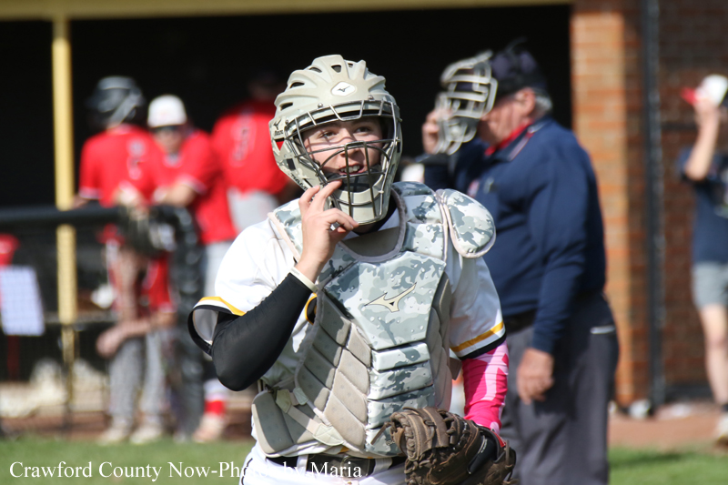 Catcher in protective gear touching his helmet on a sunny baseball field.