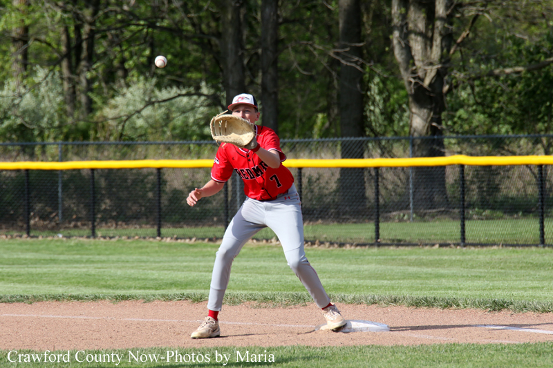 Baseball infielder in a red jersey reaches to catch a ball near first base on a sunny day.