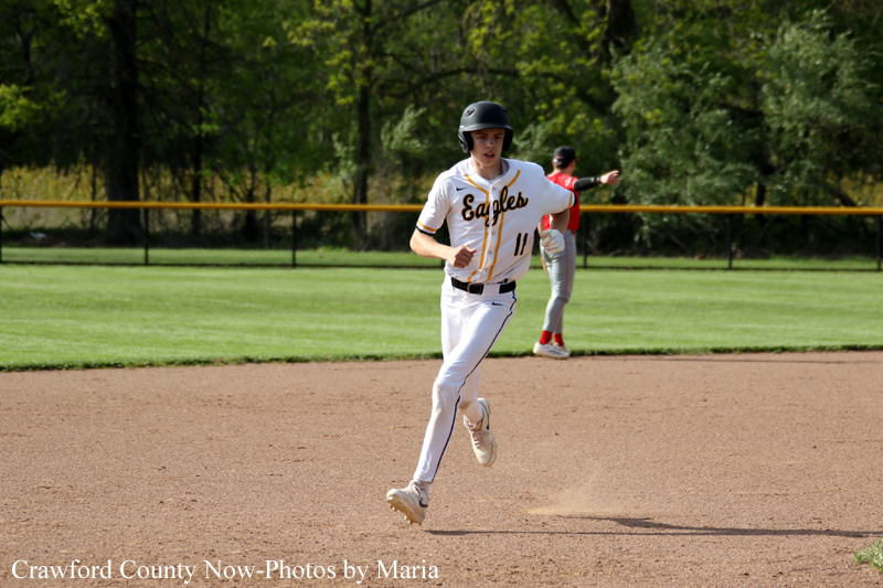 Baseball player in a white uniform runs on the dirt infield during a game, with a green outfield behind and a yellow fence in the background.