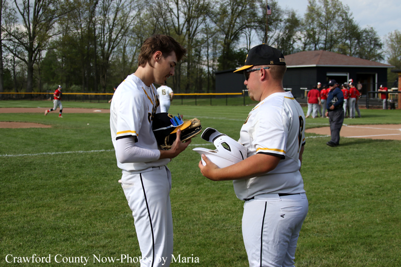 Two baseball players in white uniforms exchange gear on a grassy field; one holds a glove and the other a white helmet.