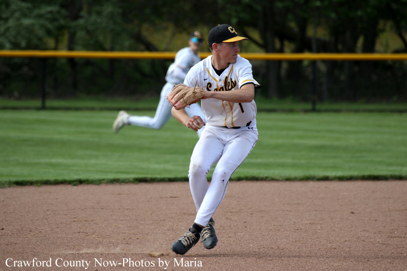 Baseball pitcher in a white and gold uniform is throwing a pitch on the infield with a teammate in the background,
