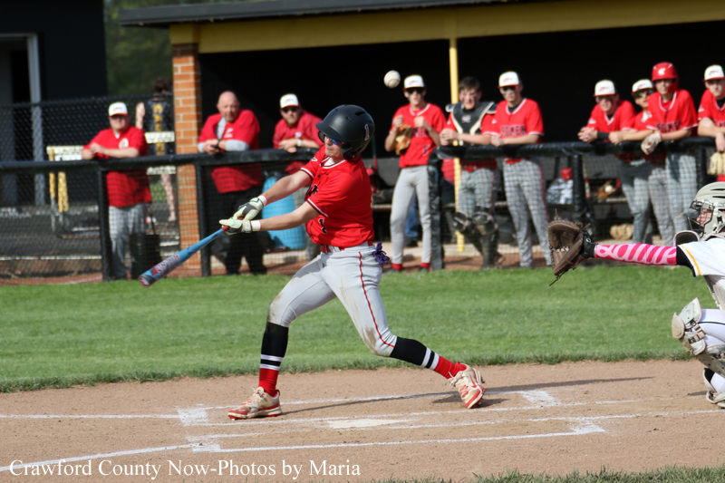 Baseball batter in red swings at a pitch as the catcher reaches for a ball in front of a dugout with spectators watching.