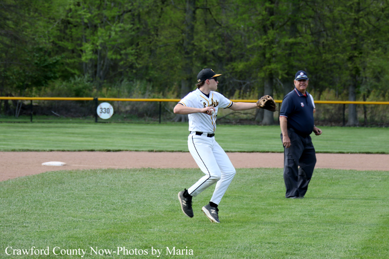 Baseball player in white uniform leaps to catch a ball on the infield, with an official standing nearby on the grass.