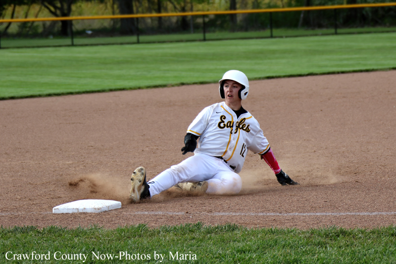 Baseball player in a white Eagles uniform slides into a base, dirt kicking up as helmeted figure reaches the bag with one arm extended.