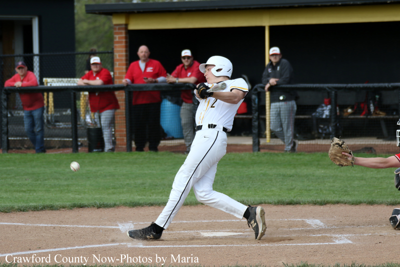 Baseball batter in a white uniform swings at a pitched ball near home plate as spectators watch from the dugout area behind the field.