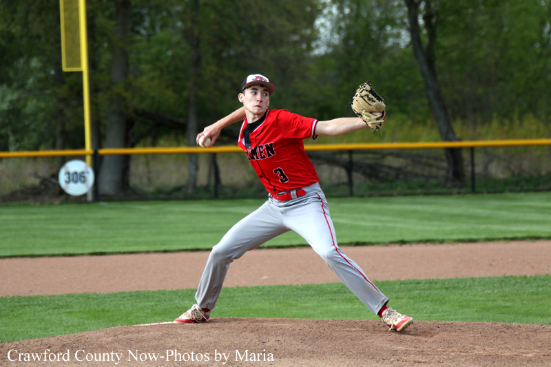 Baseball pitcher in red uniform delivers a pitch from the mound, glove extended to the left with a focused expression, in a grassy field behind a chain-link fence.