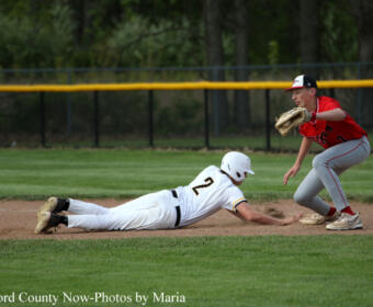 Baseball scene: a white-uniform runner slides headfirst into a dirt base while a red-uniform fielder reaches with a glove to tag him nearby on the infield fielding position.