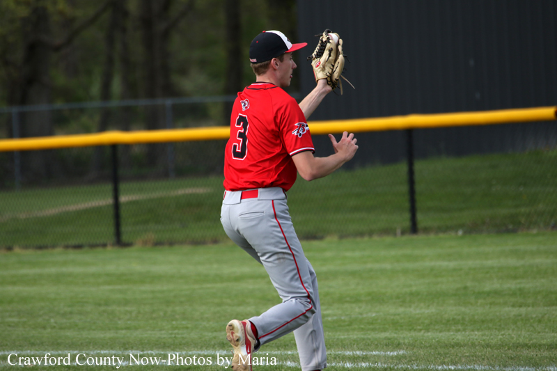 Baseball pitcher in a red jersey (number 3) mid-pitch on a grassy field with a yellow outfield fence behind him.