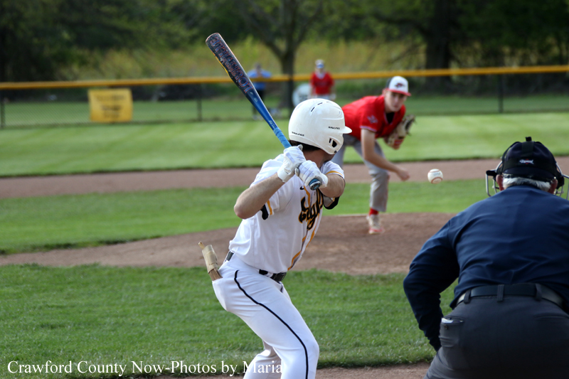 Batter in a white uniform and helmet swings at a pitch as the ball nears; an umpire in blue and a fielder in red are in the background on the baseball field.