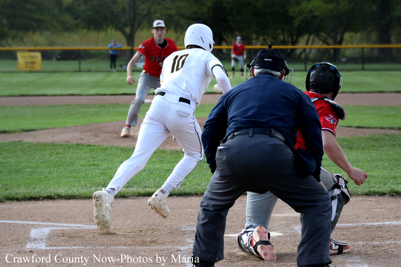 Baseball action at home plate: a baserunner in white (#10) slides toward the plate while the catcher in red blocks the run and an umpire watches nearby.
