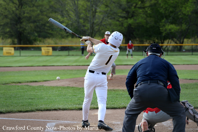 Baseball batter in white uniform swings at a pitch at home plate, with catcher and umpire in the foreground on the field.