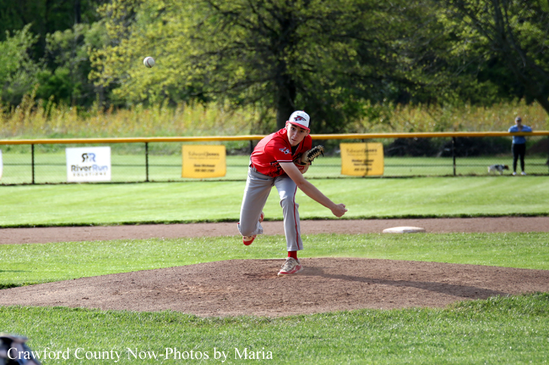 Young baseball pitcher in a red uniform delivers a pitch on the mound during a sunny game.
