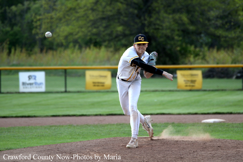 Baseball pitcher in a white uniform on the mound delivering a pitch, dirt spraying as he finishes the throw with a ball in the air to his left and outfield fencing in the background.