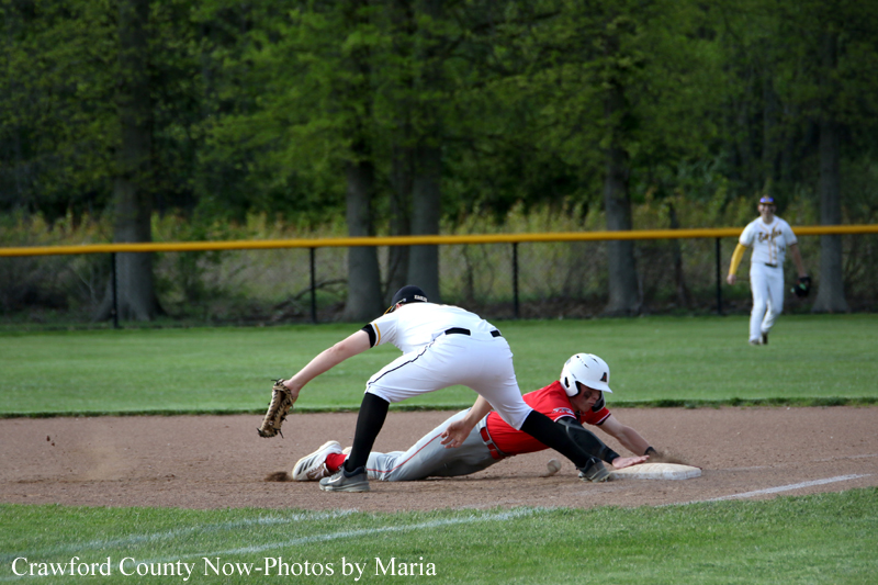 Baseball play at a bag: runner in red slides into the base as a fielder in white lunges with a glove to tag him, dust rising from the dirt. In the background, an outfielder watches on the grass field.