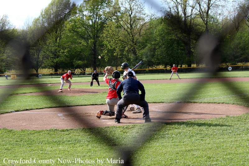 Baseball game in progress viewed through a chain-link fence, batter at plate with catcher and umpire nearby on the dirt infield.