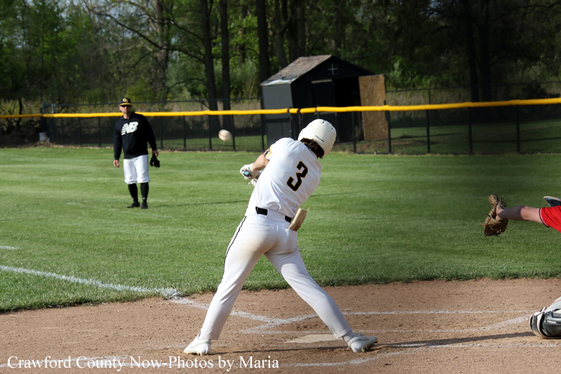 Baseball batter in white uniform #2 swings at a pitch near home plate as a fielder in black watches from the outfield on a sunny field.
